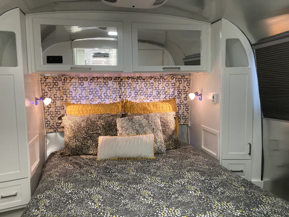 Finished Airstream bedroom with floral wall covering, overhead cabinets, and built-in bedside storage.
