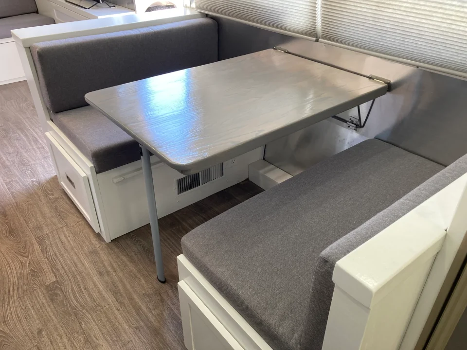 Built-in gray dinette seating around a rectangular dining table beneath the Airstream windows.