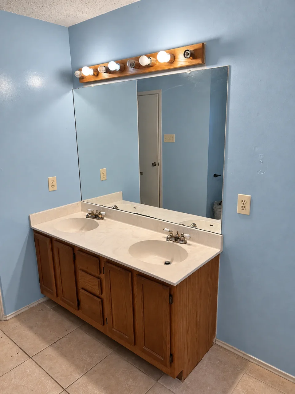 Before renovation: wood double vanity with a beige counter, wide frameless mirror and blue walls.