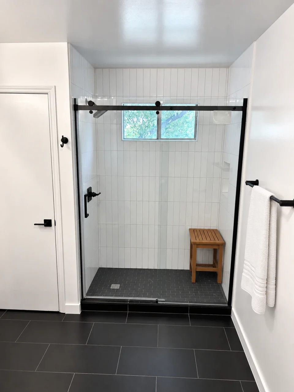 White-tiled shower with black sliding-glass hardware, a dark mosaic floor and a white towel on the adjacent rail.