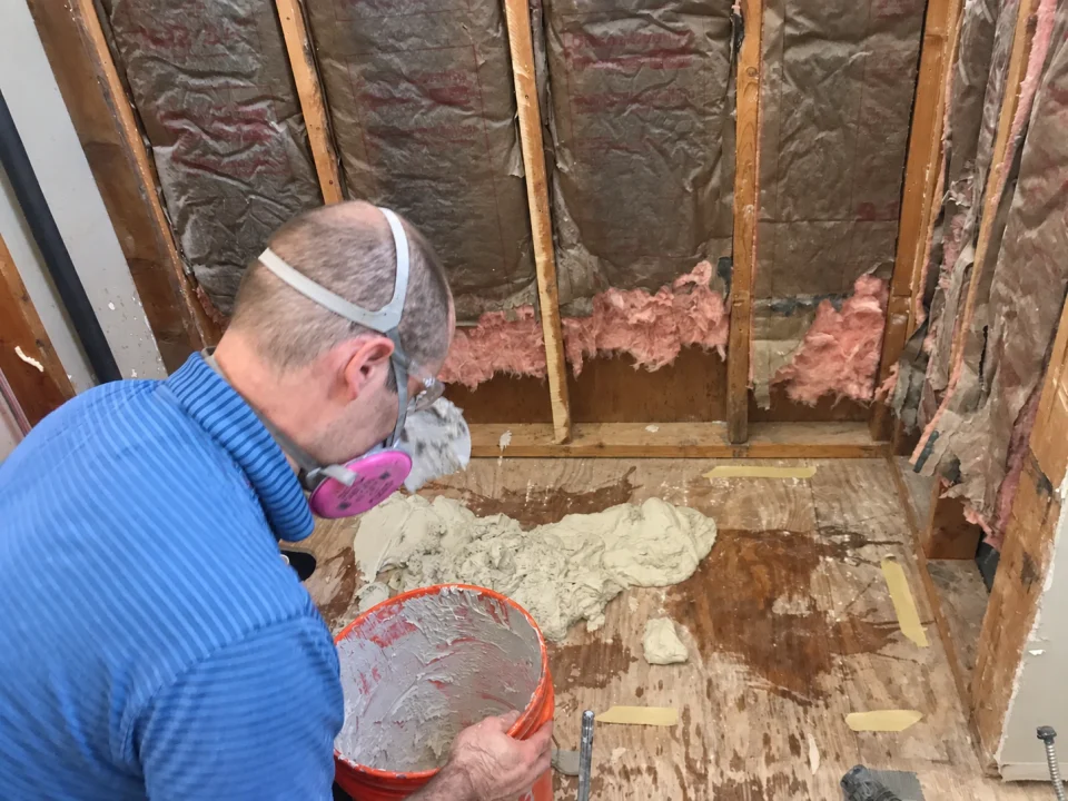 Jake working on the exposed shower floor with the wall framing and insulation visible during renovation.