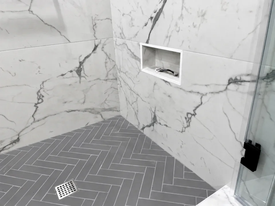 Low shaving niche in marble-look shower tile above a gray herringbone floor