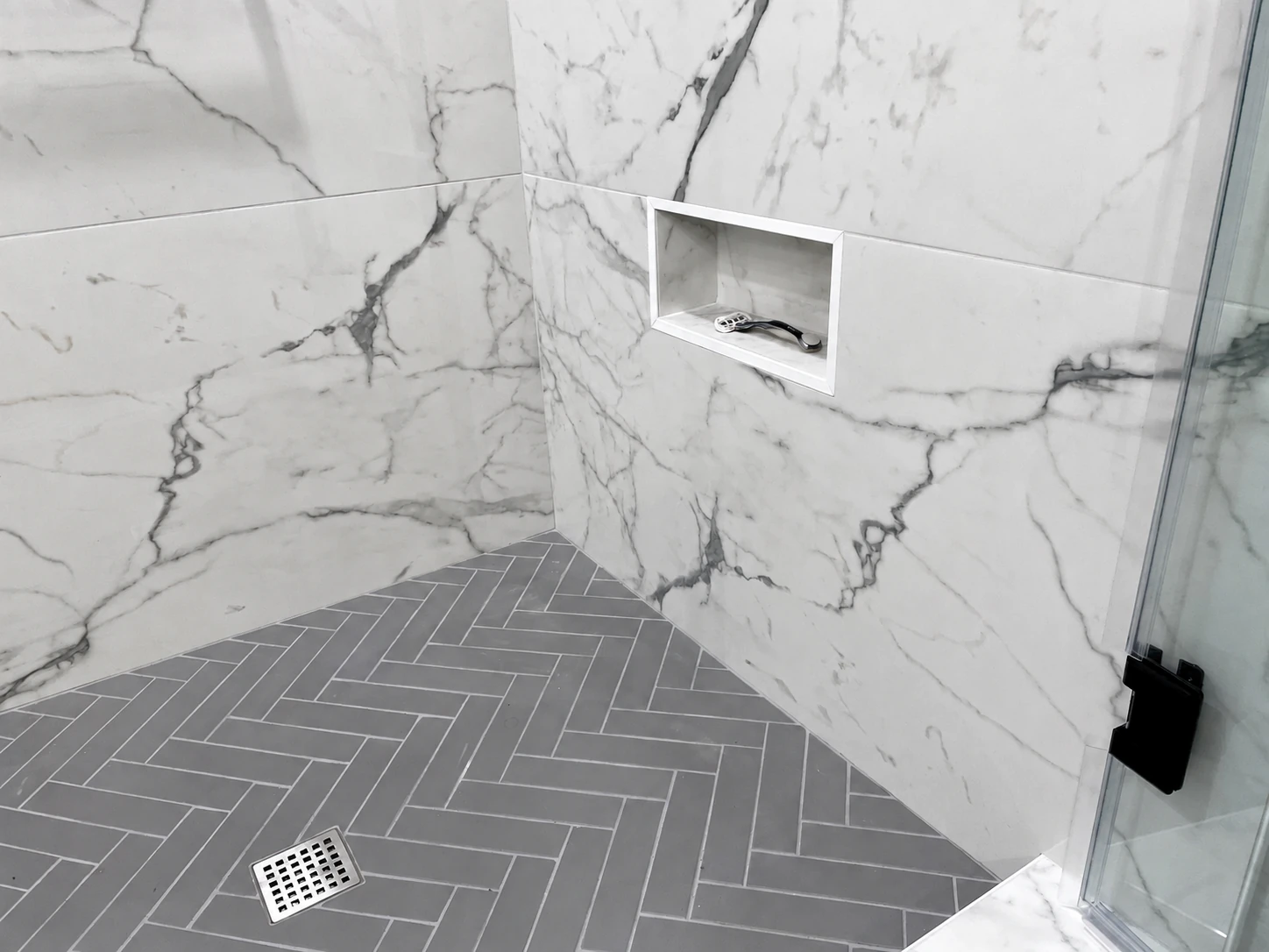 Low shaving niche in marble-look shower tile above a gray herringbone floor