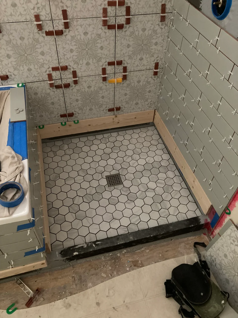 Pale sage subway tile and white patterned tile with sage and gray details being installed above a hexagonal shower floor.