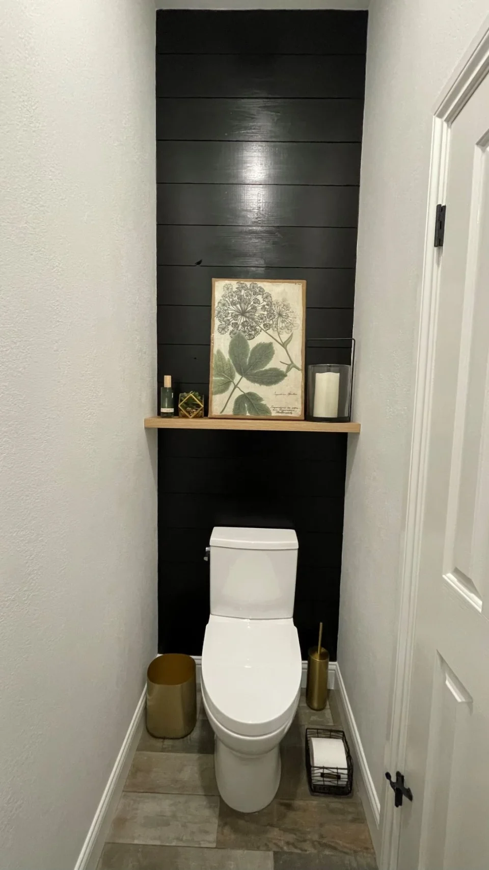 Compact powder room with white toilet, dark horizontal-board feature wall, wood shelf, and framed botanical artwork.
