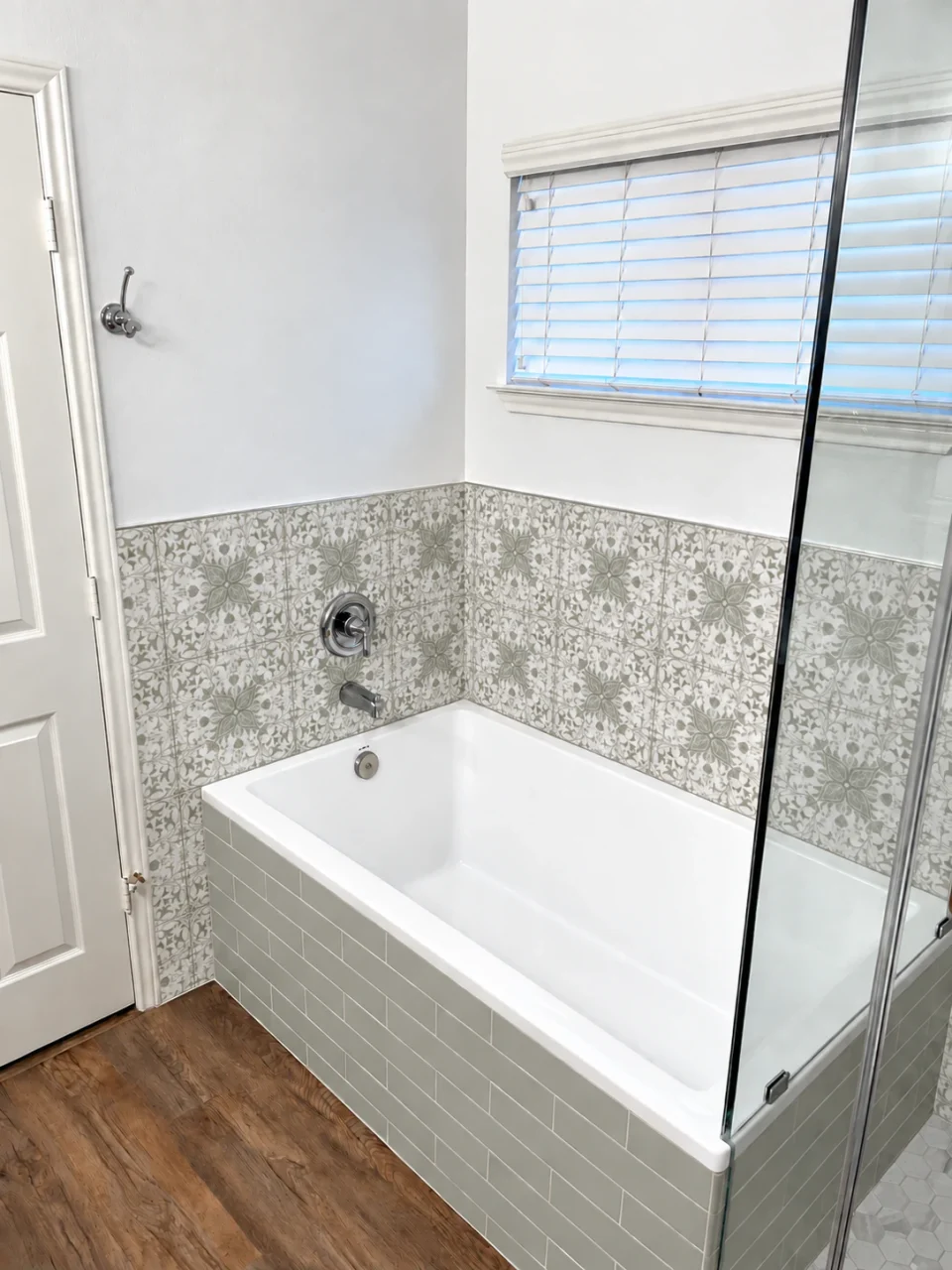 White bathtub beneath a window, with pale sage subway tile on the apron and a white patterned surround with sage and gray details.