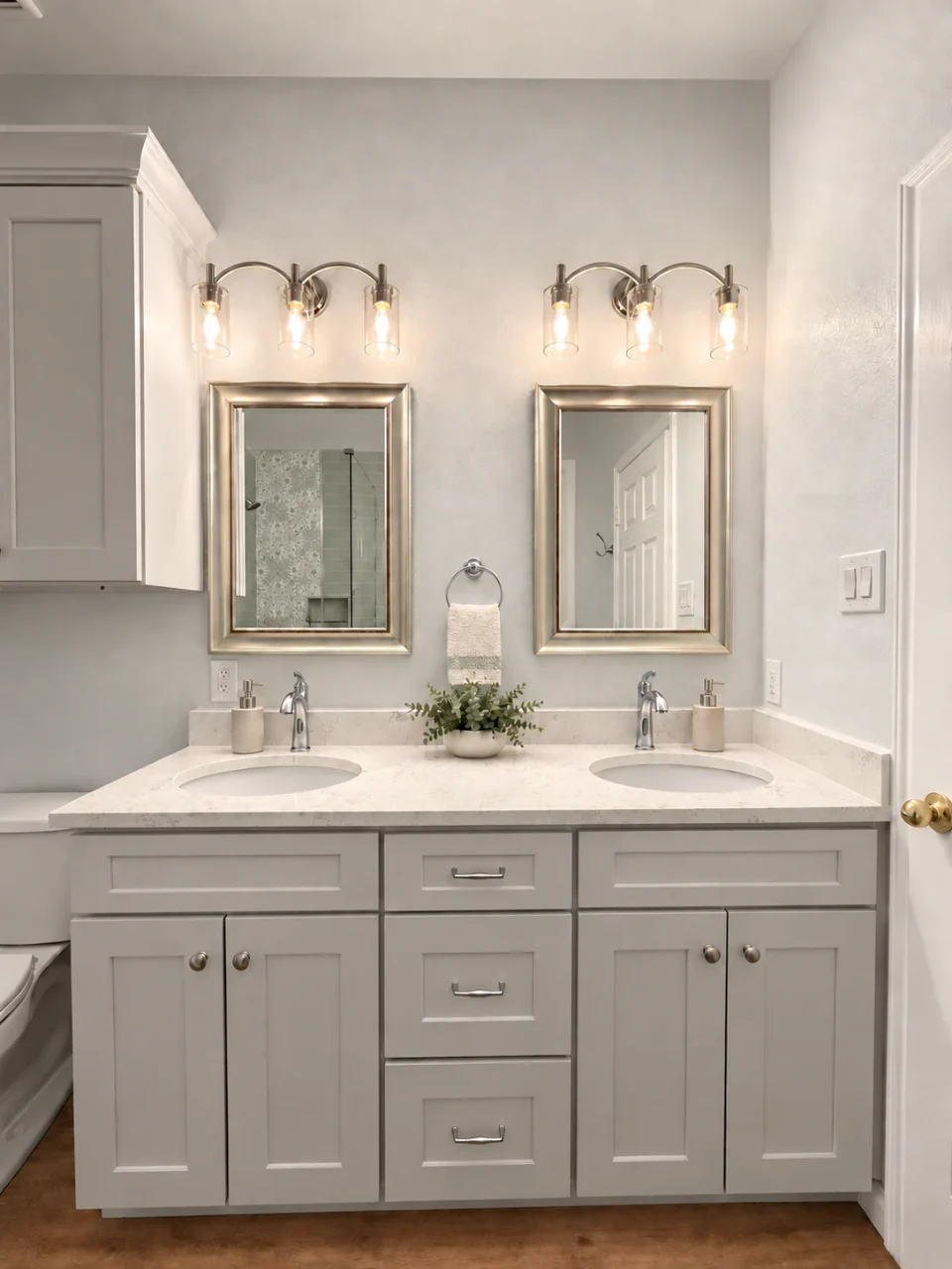 Gray double vanity with a light stone countertop, two framed mirrors, glass-shade lights and a small plant.