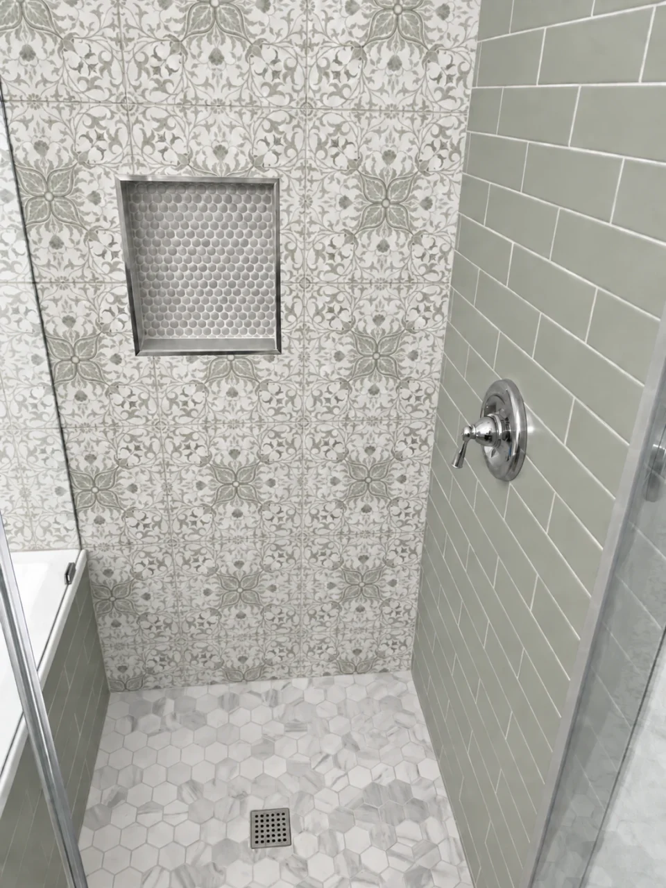 Gray penny-tile shower niche surrounded by white patterned tile with sage and gray details, beside pale sage subway tile.