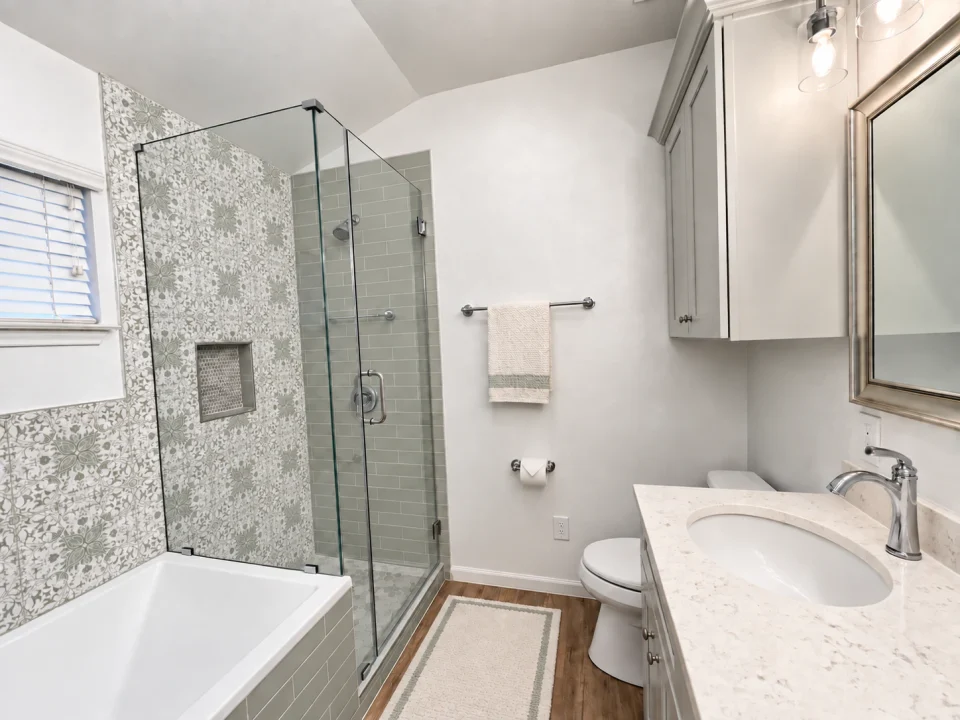 Bathroom with pale sage subway tile, white patterned tile with sage and gray details, a glass shower, white tub and gray vanity.