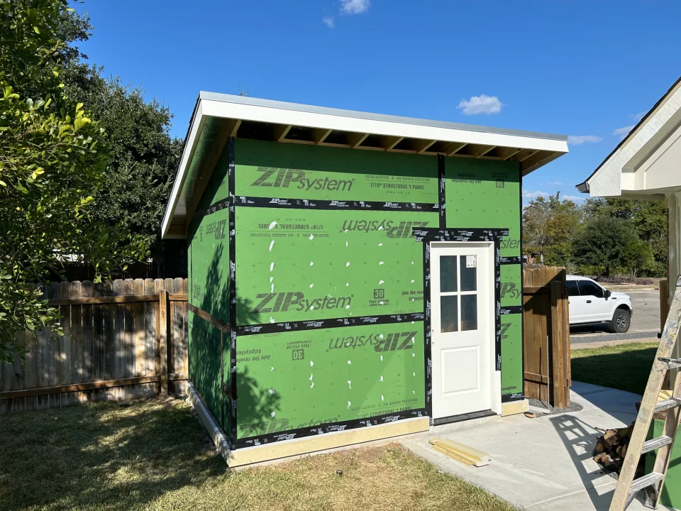 Backyard shed during construction, with green wall sheathing, exposed roof framing and a white glazed door
